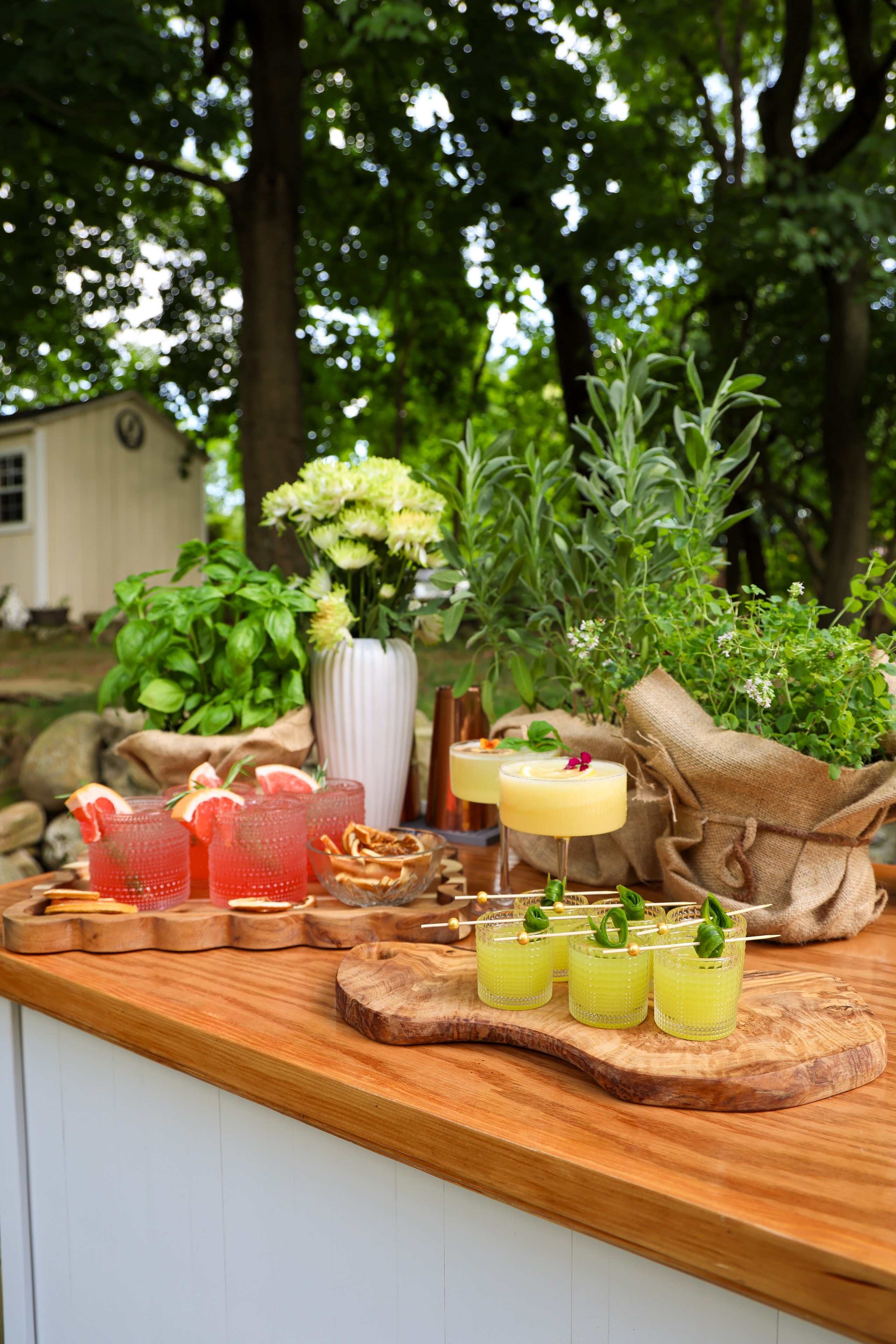 Outdoor garden bar display with colorful cocktails on wooden boards surrounded by fresh herbs